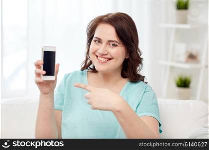 people, technology, communication and leisure concept - happy young plus size woman sitting on sofa and showing smartphone blank screen at home