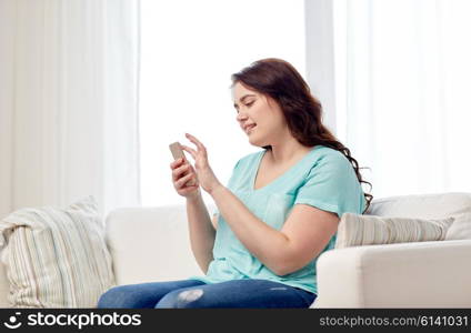 people, technology, communication and leisure concept - happy young plus size woman sitting on sofa with smartphone at home