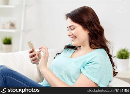 people, technology, communication and leisure concept - happy young plus size woman sitting on sofa with smartphone at home