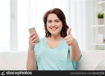 people, technology, communication and leisure concept - happy young plus size woman sitting on sofa with smartphone at home and showing thumbs up