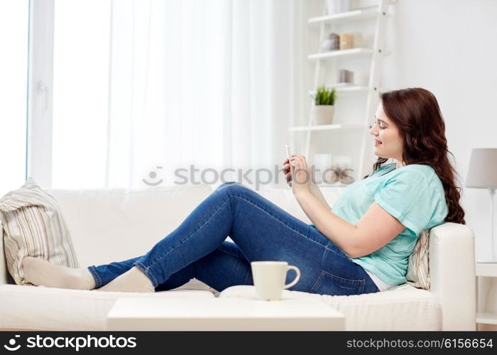 people, technology, communication and leisure concept - happy young plus size woman sitting on sofa with smartphone at home