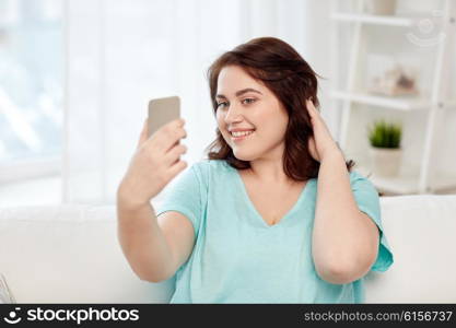 people, technology, communication and leisure concept - happy young plus size woman taking selfie with smartphone at home