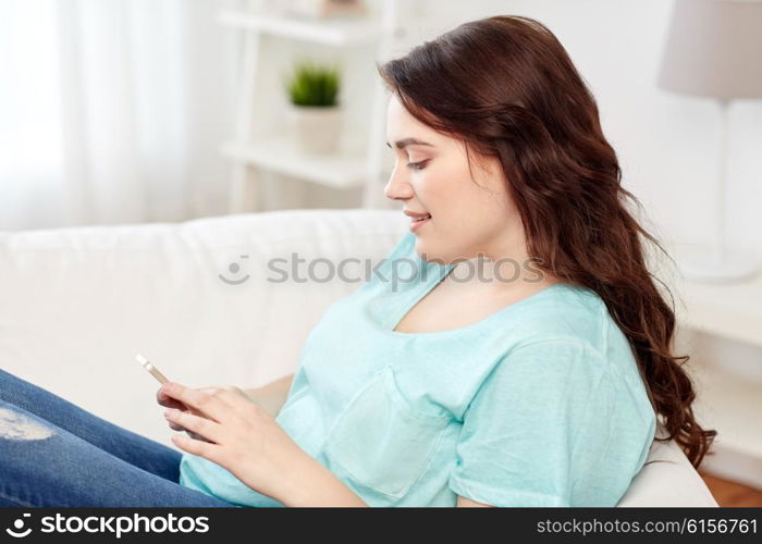 people, technology, communication and leisure concept - happy young plus size woman sitting on sofa with smartphone at home