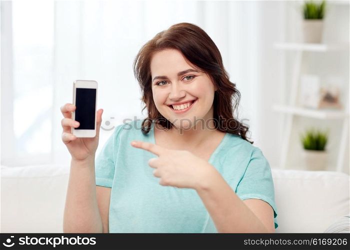 people, technology, communication and leisure concept - happy young plus size woman sitting on sofa and showing smartphone blank screen at home