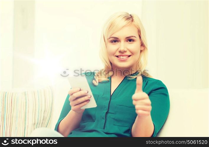 people, technology, communication and leisure concept - happy young woman sitting on sofa with smartphone at home and showing thumbs up. woman with smartphone showing thumbs up at home