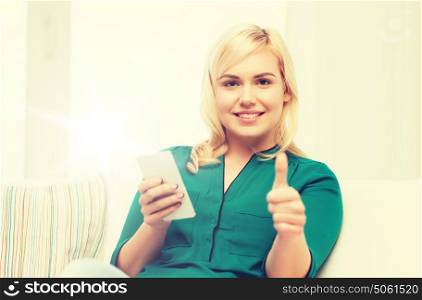 people, technology, communication and leisure concept - happy young woman sitting on sofa with smartphone at home and showing thumbs up. woman with smartphone showing thumbs up at home