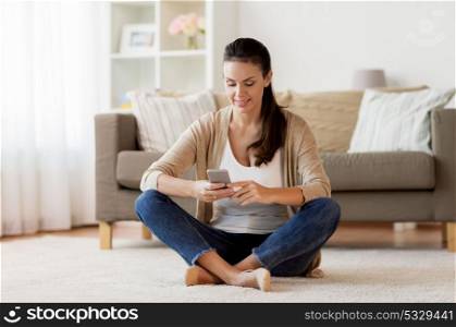 people, technology, communication and leisure concept - happy young woman sitting on floor and texting message on smartphone at home. happy woman texting message on smartphone at home