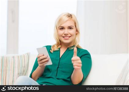 people, technology, communication and leisure concept - happy young woman sitting on sofa with smartphone at home and showing thumbs up
