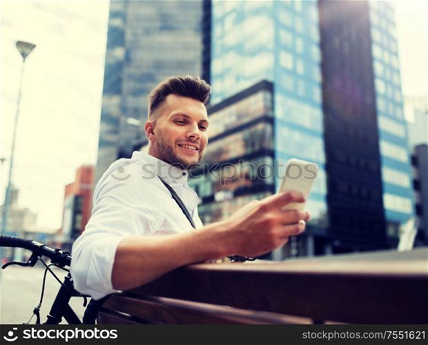 people, technology, communication and lifestyle - happy smiling young man with and bicycle sitting on city bench. happy man with smartphone and bicycle in city