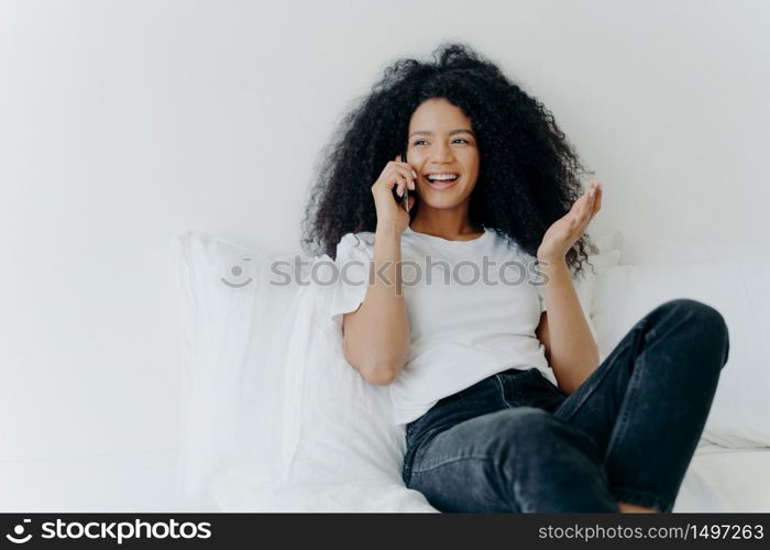 People, technology, communication and rest concept. Glad curly haired ethnic lady has telephone talk, gestures and laughs with joy, looks somewhere aside, spends time at home, sits on white bed