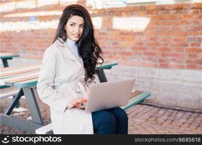 People, technology, communication concept. Beautiful female with dark wavy hair wearing white raincoat sitting at bench over brick wall holding laptop communicating online using free internet