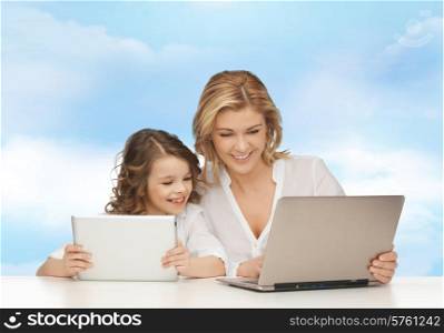 people, technology, family and parenthood concept - happy mother and daughter with laptop and tablet pc computers sitting at table over blue sky background