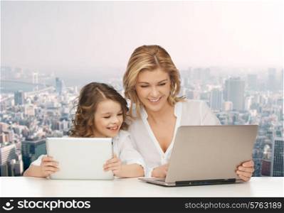 people, technology, family and parenthood concept - happy mother and daughter with laptop and tablet pc computers sitting at table over city background