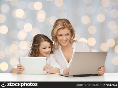 people, technology, family and parenthood concept - happy mother and daughter with laptop and tablet pc computers sitting at table over holidays lights background