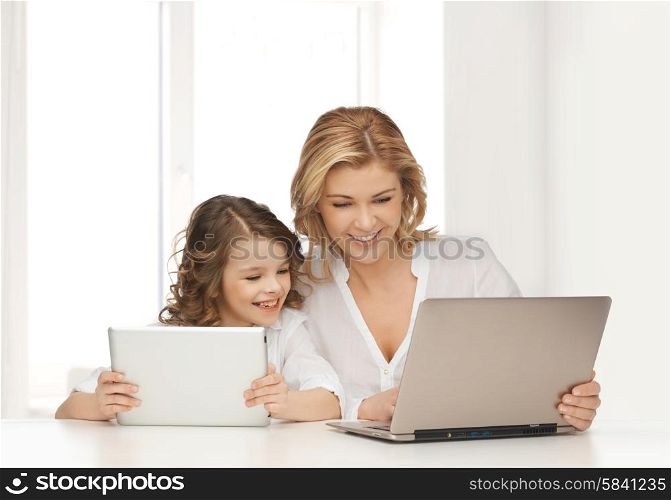 people, technology, family and parenthood concept - happy mother and daughter with laptop and tablet pc computers sitting at table over white room background