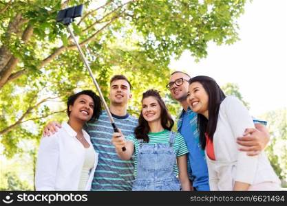 people, technology, friendship and international concept - group of happy friends or students taking picture by smartpnone on selfie stick at summer park. happy friends taking photo by selfie stick at park