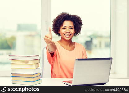 people, technology, gesture and education concept - happy african american young woman sitting at table with laptop computer and books showing thumbs up at home