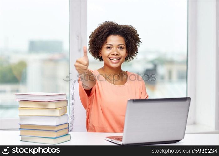 people, technology, gesture and education concept - happy african american young woman sitting at table with laptop computer and books showing thumbs up at home