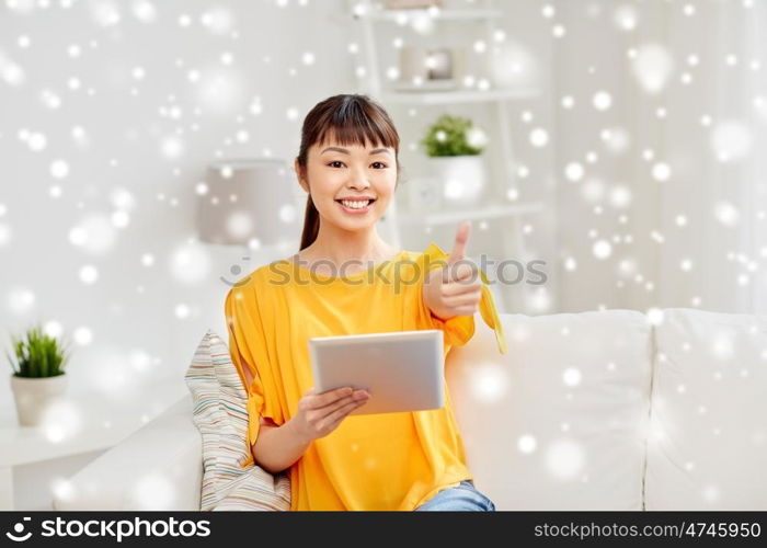 people, technology, gesture and leisure concept - happy young asian woman sitting on sofa with tablet pc computer showing thumbs up at home over snow