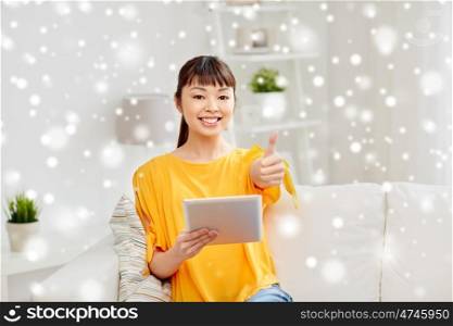people, technology, gesture and leisure concept - happy young asian woman sitting on sofa with tablet pc computer showing thumbs up at home over snow