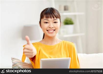 people, technology, gesture and leisure concept - happy young asian woman sitting on sofa with tablet pc computer showing thumbs up at home