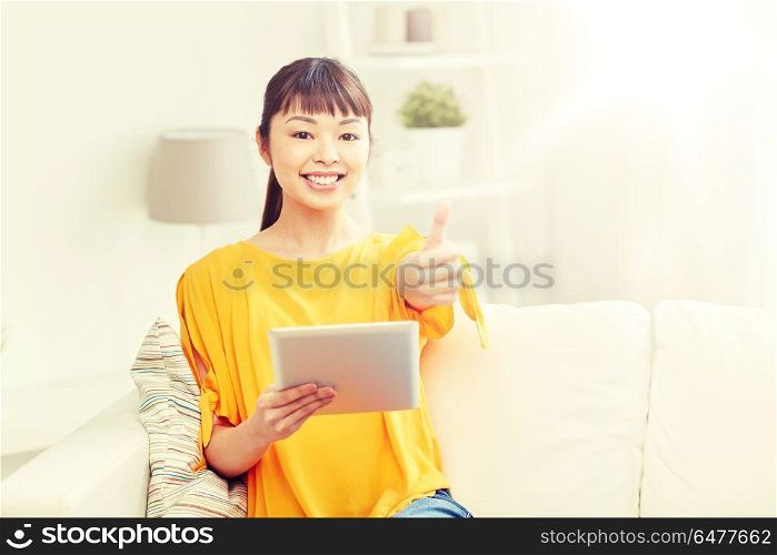 people, technology, gesture and leisure concept - happy young asian woman sitting on sofa with tablet pc computer showing thumbs up at home. happy asian woman with tablet pc and thumbs up. happy asian woman with tablet pc and thumbs up