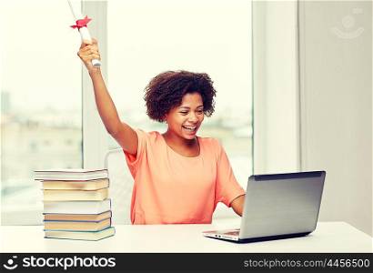 people, technology, graduation and education concept - happy african american young woman sitting at table with laptop computer, books and diploma at home