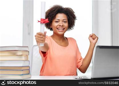people, technology, graduation and education concept - happy african american young woman sitting at table with laptop computer, books and diploma at home