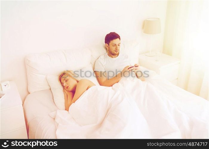 people, technology, internet and communication concept - man with smartphone texting message while woman is sleeping in bed