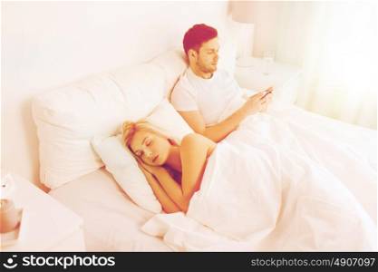 people, technology, internet and communication concept - man with smartphone texting message while woman is sleeping in bed. man texting message while woman is sleeping in bed