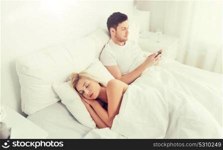 people, technology, internet and communication concept - man with smartphone texting message while woman is sleeping in bed. man texting message while woman is sleeping in bed
