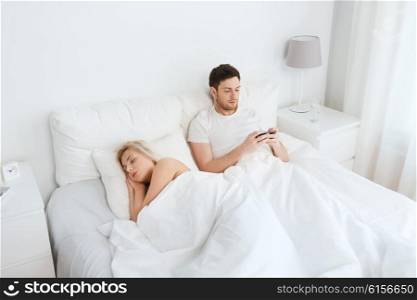 people, technology, internet and communication concept - man with smartphone texting message while woman is sleeping in bed