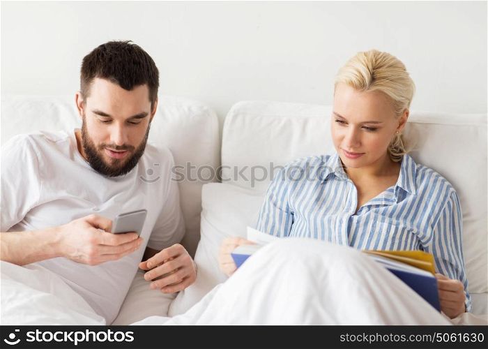 people, technology, internet, relations and communication concept - happy couple with book and smartphones in bed at home. couple with book and smartphones in bed at home