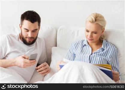 people, technology, internet, relations and communication concept - happy couple with book and smartphones in bed at home. couple with book and smartphones in bed at home