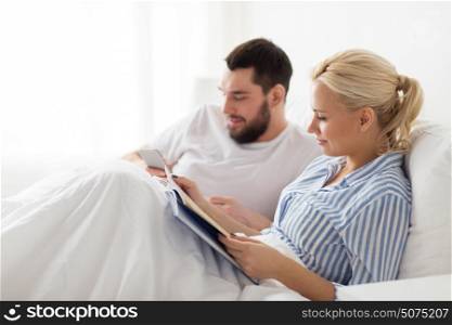 people, technology, internet, relations and communication concept - happy couple with book and smartphone in bed at home. couple with book and smartphone in bed at home