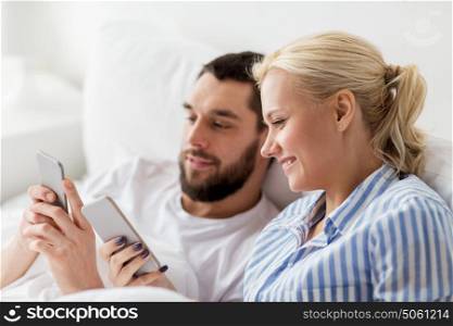 people, technology, internet, relations and communication concept - happy couple with smartphones in bed at home. happy couple with smartphones in bed at home