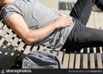 people, technology, leisure and lifestyle - close up of man with smartphone, earphones and backpack listening to music lying on city street bench