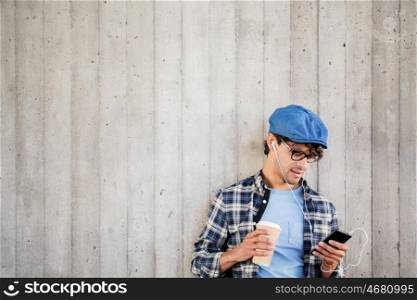 people, technology, leisure and lifestyle - man with earphones and smartphone drinking coffee and listening to music at street wall