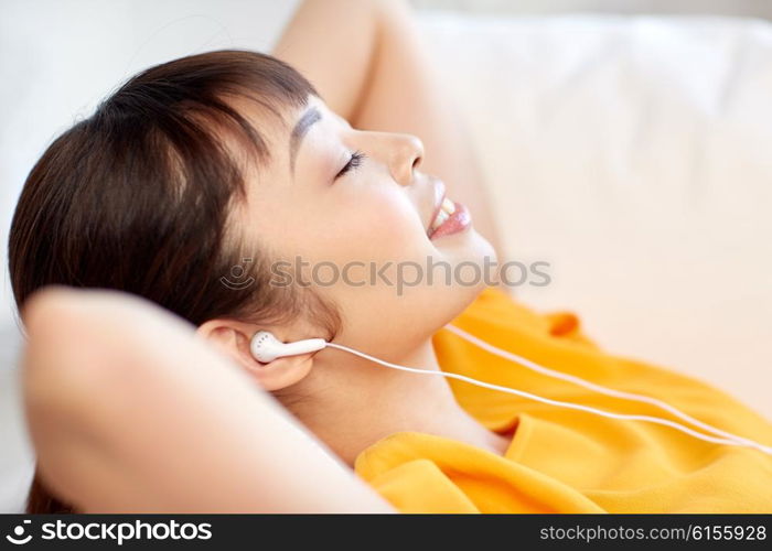 people, technology, relax and leisure concept - happy asian young woman lying on sofa with earphones listening to music at home