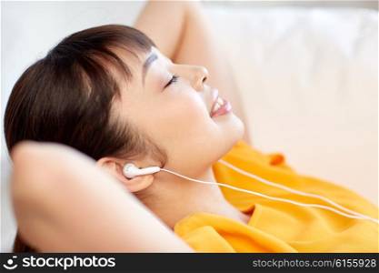 people, technology, relax and leisure concept - happy asian young woman lying on sofa with earphones listening to music at home