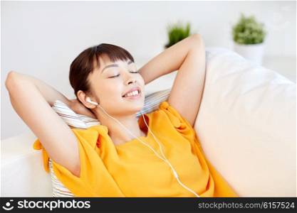 people, technology, relax and leisure concept - happy asian young woman lying on sofa with earphones listening to music at home