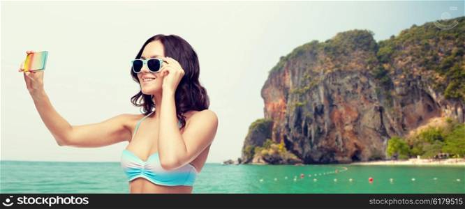 people, technology, summer and beach concept - happy young woman in bikini swimsuit and sunglasses taking selfie with smatphone over sea and rock background