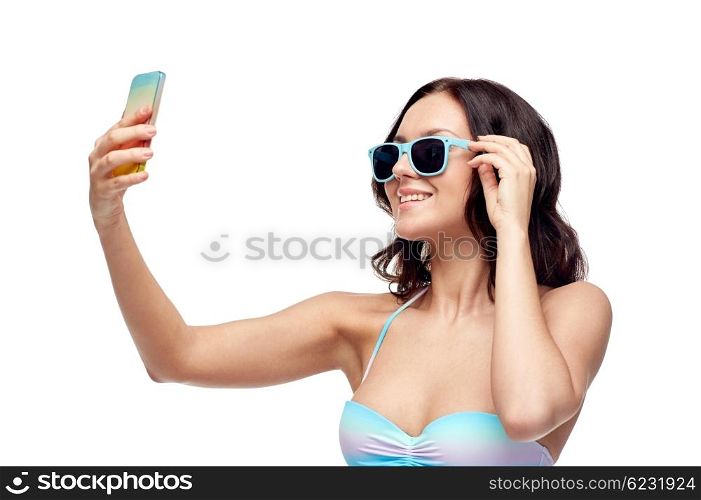 people, technology, summer and beach concept - happy young woman in bikini swimsuit and sunglasses taking selfie with smatphone
