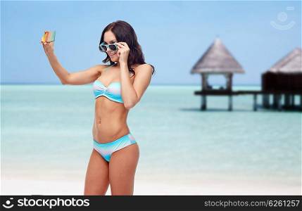 people, technology, summer and travel concept - happy young woman in bikini swimsuit and sunglasses taking selfie with smatphone over maldives beach with bungalow background