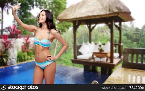 people, technology, summer holidays, travel and tourism concept - happy young woman in bikini swimsuit taking selfie with smatphone over hotel resort background