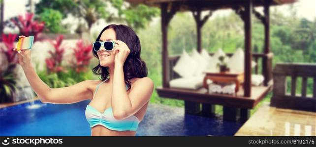 people, technology, summer holidays, travel and tourism concept - happy young woman in bikini swimsuit and sunglasses taking selfie with smatphone over hotel resort background