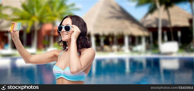 people, technology, summer vacation and travel concept -young woman in bikini swimsuit and sunglasses taking selfie with smatphone over swimming pool, bungalow at hotel resort background