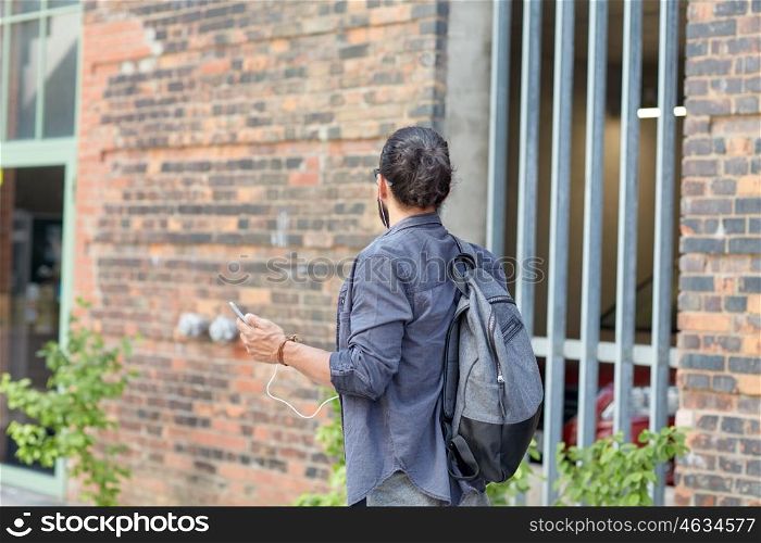 people, technology, travel and tourism - man with backpack, smartphone and earphones walking along city street and listening to music