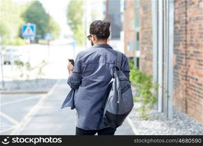people, technology, travel and tourism - man with backpack, smartphone and earphones walking along city street and listening to music