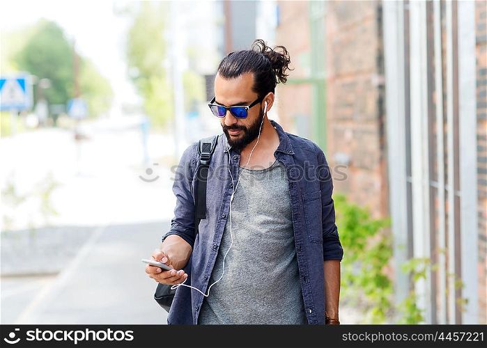 people, technology, travel and tourism - man with earphones, smartphone and bag walking along city street and listening to music